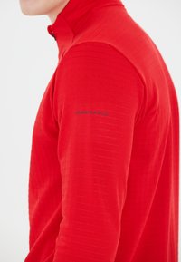 Rode langemouwen top met hoge kraag, voorzien van een horizontale ribbeltextuur en een zwart "ENDURANCE" logo op de linker mouw.