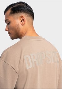 T-shirt oversize beige avec un col rond et le texte "DROPSIZE" imprimé au dos dans une teinte légèrement plus foncée. Texture douce.