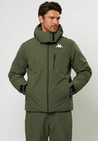 Egy férfi olívazöld kapucnis télikabátot visel, cipzáras zsebekkel és egy fehér logóval a mellkasán, kezét a zsebben tartja, és jobbra néz.