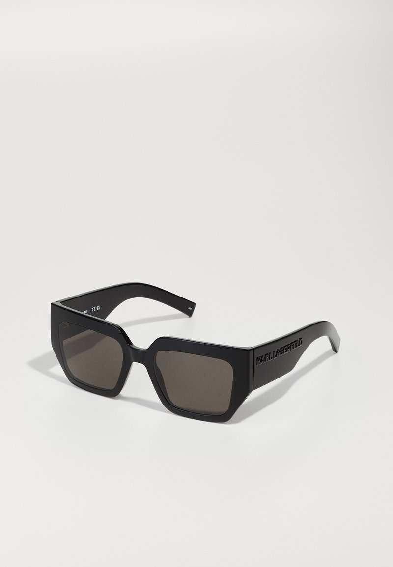 Lunettes de soleil noires rectangulaires avec verres foncés, monture épaisse et détails de logo sur les branches. Design angulaire avec texture lisse.