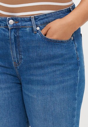 Personne portant un jean en denim bleu avec une main placée dans la poche avant, portant un haut marron et blanc à rayures.