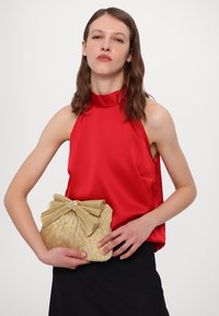 Haut à bretelles rouge, pochette plissée dorée avec un accent en nœud, jupe noire. La pochette présente une surface texturée et est tenue dans les mains du modèle.