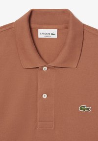 Brun polo-tröja i texturerad tyg, med treknappsslutning, ribbad krage och en liten grön krokodil-logotyp på bröstet.