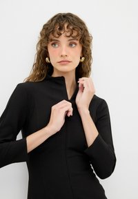 Schwarzes Reißverschlusskleid mit hohem Kragen, Dreiviertelärmeln und glatter Textur. Das Model zieht den Reißverschluss hoch und trägt goldene Creolen.