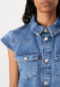 Leren denimvest in lichtblauw. Heeft een kraag, kapmouwen en twee borstzakken met zilverkleurige knopen. Verweerde textuuraccenten.