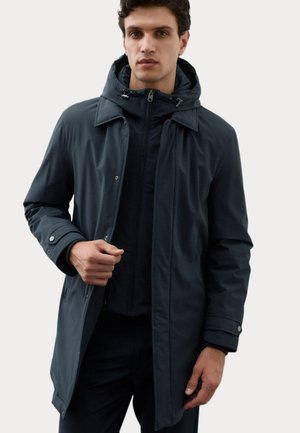 Homme portant un manteau à capuche foncé par-dessus une veste zippée, tenant le manteau ouvert d'une main devant un fond clair uni.