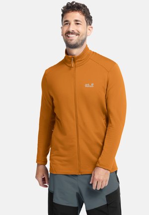 Mann lächelt, trägt eine orangefarbene Jacke mit Reißverschluss und hohem Kragen sowie graue und schwarze Hosen vor einem schlichten weißen Hintergrund.