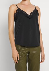 Camisola in satin nero con scollatura a V, rifinita con pizzo lungo i bordi e spalline sottili e regolabili. Indossata con pantaloni aderenti verde oliva.