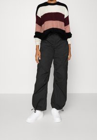 Pull en maille épaisse avec des rayures noires, bordeaux, rose pâle et blanches. Associé à un pantalon cargo noir ample et des baskets blanches.