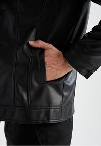 Veste en cuir noir avec une poche latérale, texture lisse et couture détaillée le long de l'avant et de l'ourlet. Les poignets présentent des accents contrastants.