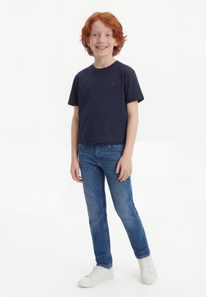 Rudy chłopiec uśmiechający się, ubrany w granatowy t-shirt, niebieskie dżinsy i białe trampki, stojący na białym tle.