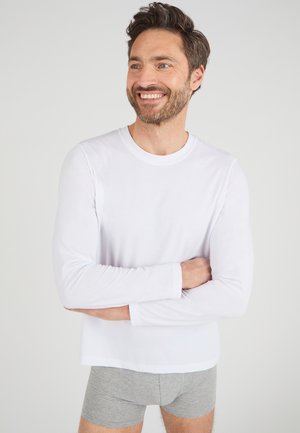 Chemise blanche à manches longues en matière douce, encolure ronde, avec une coupe classique. Portée avec un short en coton gris, les bras croisés devant.