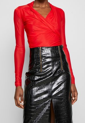 Frau trägt ein langärmeliges rotes Wickeloberteil und einen hoch taillierten schwarzen Rock aus Kunstkrokodilleder mit Reißverschlüssen vorne und einem Schlitz vorne.