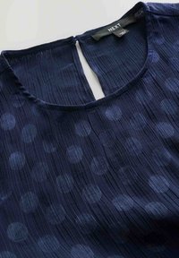 Marineblaue Bluse mit rundem Ausschnitt, strukturierten Falten und einem Tupfenmuster. Rückenverschluss mit einem Schlitzdetail am Nacken.