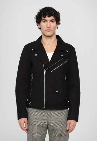 Veste de moto noire en tissu lisse, avec un col cranté, une fermeture éclair asymétrique et plusieurs poches zippées. Accompagnée de pantalons à carreaux.