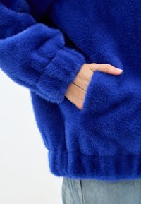 Giacca in pelliccia sintetica blu con una finitura morbida e texturizzata. Caratterizzata da polsini a coste e una tasca frontale, offre calore e comfort.