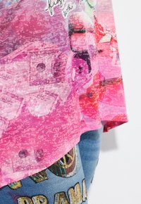 Camisa gráfica de manga corta rosa con texturas en capas, que presenta patrones de dinero y elementos coloridos; llevada sobre vaqueros de denim adornados.
