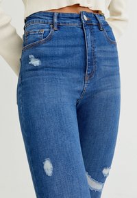 Högmidjade blå jeans i denim, med slitna detaljer, fransade kanter och ljusa fläckar på framsidan.