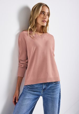 Lyserød, strikket sweater med V-hals, ribbede manchetter og kant, parret med blå jeans. Stoffet ser blødt og afslappet ud.