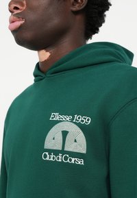 Grøn hoodie med et hvidt broderet logo med geometrisk design, "Ellesse 1959" og "Club di Corsa" tekst på brystområdet.