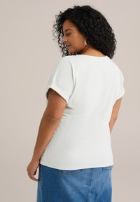 Top bianco texturizzato con maniche corte arrotolate, visto da dietro, abbinato a una gonna di jeans. Presenta una vestibilità rilassata e un tessuto morbido.