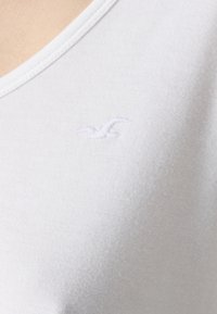 Bela bombažna majica z V-izrezom in subtilnim izvezenim logotipom ptice v svetlo sivi barvi na predelu prsnega koša. Gladka tekstura.