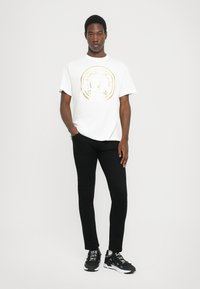 Λευκό βαμβακερό μπλουζάκι με χρυσό κυκλικό λογότυπο, συνδυασμένο με μαύρα τζιν στενού κοψίματος και μαύρα αθλητικά παπούτσια με λευκές λεπτομέρειες και κορδόνια.