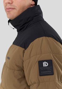 Padded jacket featuring a black upper section and brown lower body. Includes a logo patch on the left sleeve and adjustable collar.