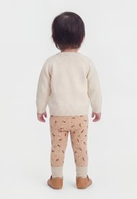 Batole stojící zády, oblečené v krémovém pleteném svetru, béžových legínách s malým červeným květinovým vzorem a hnědých botách.