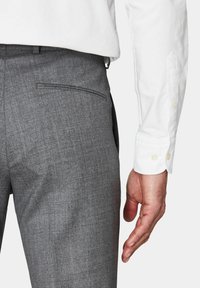 Pantaloni grigio carbone con tessuto testurizzato e una sola tasca sul retro. È visibile una camicia bianca a maniche lunghe, con dettagli dei bottoni sul polsino.