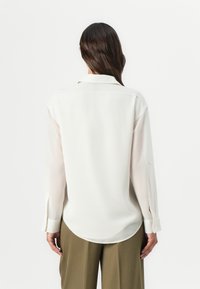 Lightweight, cream blouse with a collared neckline, long sleeves, and a rounded hem. Features buttoned cuffs and a smooth texture.