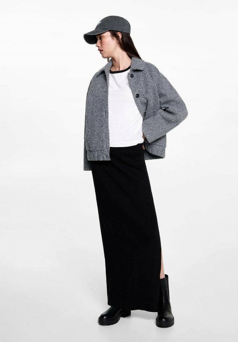 Šedá vlněná bunda s knoflíkovým zapínáním, bílý tričko, dlouhá černá žebrovaná sukně a černé kotníkové boty, doplněné šedou kšiltovkou.