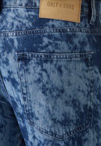 Jeans de mezclilla azul claro con un patrón desvaído y moteado. Presenta una etiqueta de cuero en la parte posterior, bolsillos cosidos y una textura ligeramente desgastada.