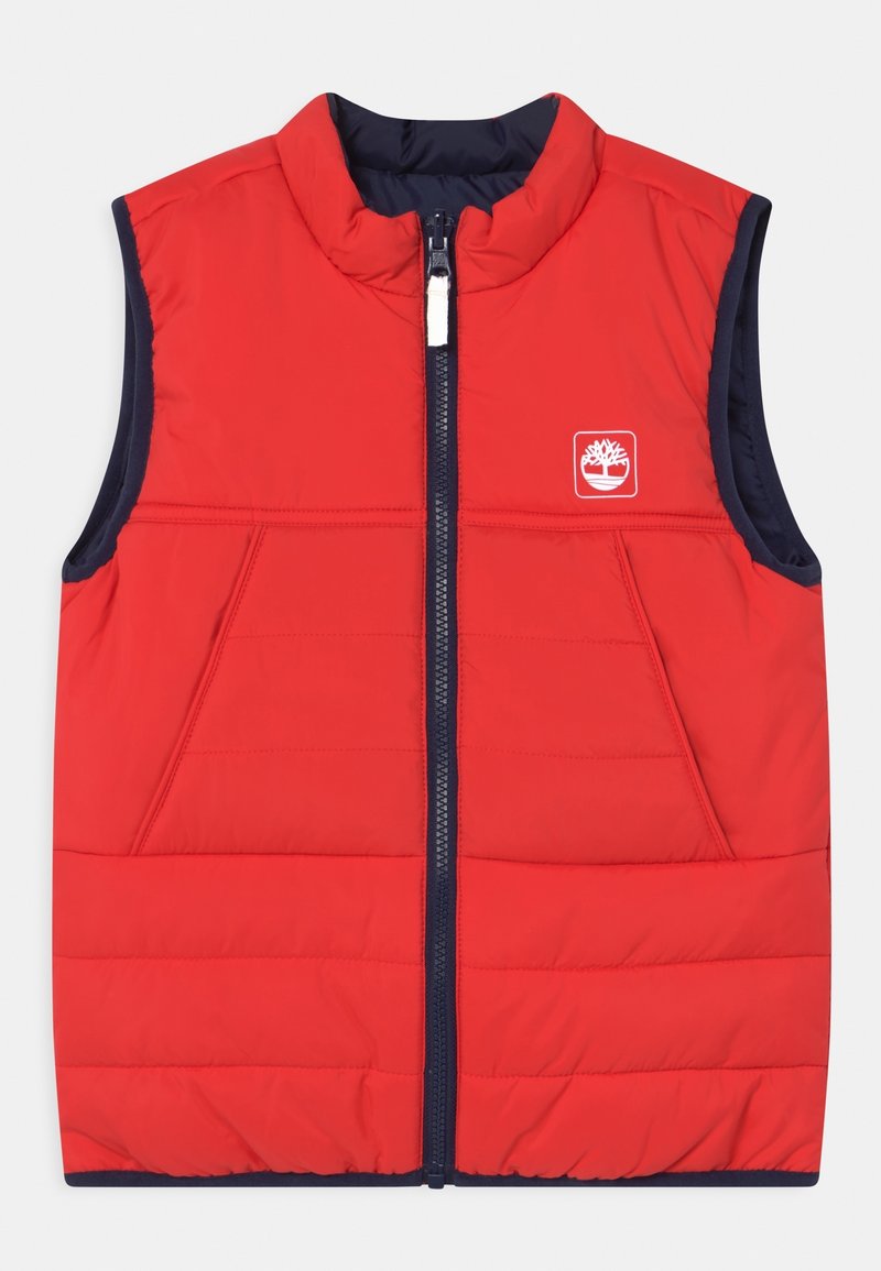 Veste sans manches matelassée rouge avec fermeture éclair frontale, bordure bleu marine et logo blanc sur le haut gauche de la poitrine.