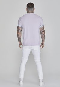 Gaiši purpura t-krekls ar baltām uzrakstiem uz piedurknēm, kombinēts ar pieguļošām baltām biksēm un baltām sporta kedām. Aizmugures skats parāda tetovējumu detaļas.