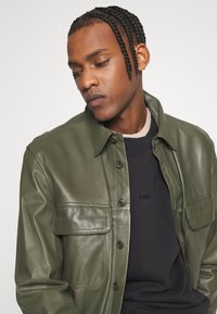 Giacca di pelle verde oliva con chiusura a bottoni, due tasche sul petto e texture liscia, indossata sopra un maglione nero a girocollo.