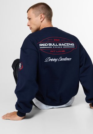 Junger Mann mit kurzen Haaren sitzt mit dem Rücken zur Kamera, trägt einen marineblauen Pullover mit dem Text "Red Bull Racing Pit Lane Driving Excellence" auf dem Rücken.