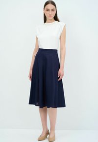 Top bianco a maniche corte con scollatura decorativa abbinato a una gonna svasata blu scuro. Il modello indossa dei tacchi beige, mostrando un tessuto liscio e una vestibilità strutturata.