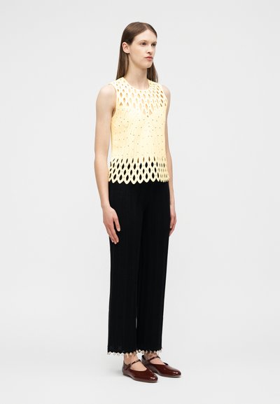 Femme portant un haut sans manches jaune découpé au laser, un pantalon large noir avec des ourlets perlés, et des chaussures plates Mary Jane marron, debout devant un fond uni.