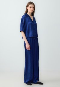 Navyblauwe tweedelige outfit van lichtgewicht stof; bestaat uit een kortomslagen overhemd met knopen en losse, verstelbare broek.