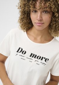 Tricou din bumbac de culoare crem, cu text negru care spune „Faceți mai mult din ceea ce vă face fericiți. PROTEJAȚI-VĂ PACEA.” Cu guler rotund și mâneci scurte.