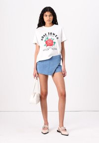 T-shirt blanc avec un motif "Rose Power" et un imprimé de rose rouge, associé à une jupe portefeuille en denim bleu, un sac blanc et des chaussures claires.