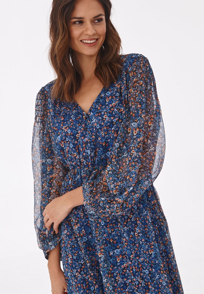 Femme souriante, portant une robe à fleurs bleues à manches longues avec des manches bouffantes transparentes, se tenant devant un fond blanc.