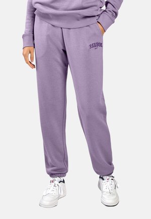 Persoon die paarse Reebok joggingbroek en bijpassende trui draagt, met witte Reebok-sneakers, staand met één hand in de zak.