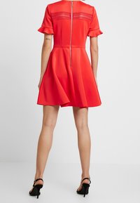 Robe rouge avec des manches courtes à volants, en tissu lisse, et une fermeture éclair au dos ; jupe évasée, longueur genoux, texture simple. Talons noirs.