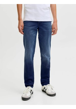 Person iført blå jeans, hvid t-shirt og hvide sneakers med sorte striber, der står på en ensfarvet lys baggrund.