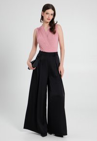 Top senza maniche di un rosa polveroso con pieghe e brillanti abbinato a pantaloni neri a vita alta con gamba larga. Borsa a tracolla nera in mano.