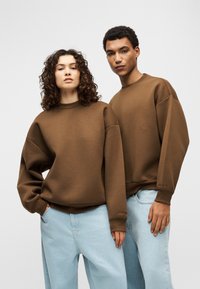 Καφέ oversized φούτερ με χαμηλωμένους ώμους, ριμπ μανσέτες και απαλή υφή, συνδυασμένα με ανοιχτό μπλε τζιν παντελόνια σε χαλαρή γραμμή.