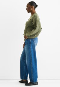 Grüne Strickjacke mit strukturierten Noppen, kombiniert mit weit geschnittenen blauen Jeans und schwarzen Ballerinas. Drei-Viertel-Profilansicht vor einem weißen Hintergrund.