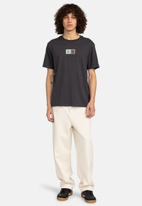 Dunkelgraues Baumwoll-T-Shirt mit kurzen Ärmeln, versehen mit einem zentralen Grafikdesign aus mintgrünen und grauen Quadraten. Kombiniert mit cremefarbenen Hosen und schwarzen Sneakern.